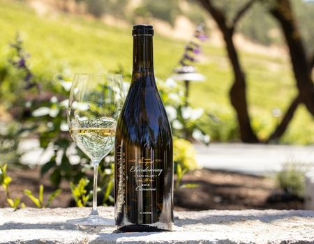 A bottle of Frank Family's Lewis Vineyard Chardonnay and wine glass set against a vineyard