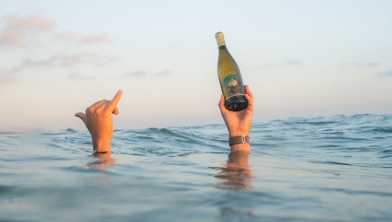 Two hands splashing out of the ocean holding a bottle of Carneros Chardonnay and wearing the custom 4ocean bracelet